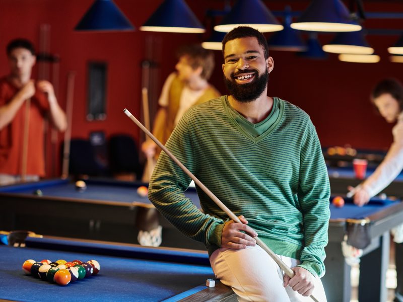 A man smiles while holding his pool cue, enjoying a casual game of billiards with his friends.